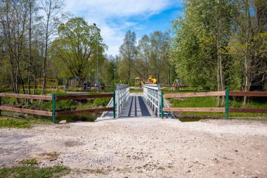 Metal korkuluklu ve ahşap aksanlı küçük bir köprü dereyi geçer, parka götürür, ağaçlarla ve açık alanlarla çevrili..