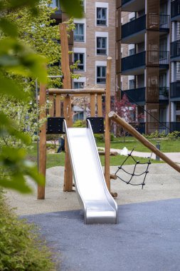 Metal kaydırak, ahşap çerçeve ve yollar, yeşillikler ve çok katlı apartmanlarla çevrili tırmanma ağıyla modern oyun alanı.