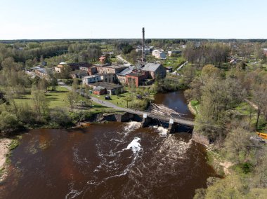 Eski tuğla binalar, uzun bir baca, beyaz suyu olan bir nehir barajı, dağınık ağaçlar ve yapılar olan sanayi bölgesinin gelişmiş perspektifi..