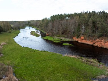Yeşil çimlerle çevrili dolambaçlı bir nehir ve sağ tarafında kırmızı turuncu kayalıkların yanı sıra yüksek ağaçlarla çevrili ormanlık alanlar..