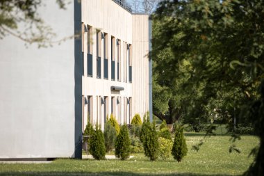 Açık renkli cepheli ve uzun pencereli modern bir dikdörtgen bina, kısmen ağaçlarla çevrili ve bir çimenlik ve küçük çalılarla çevrili..