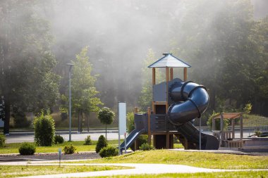Sarmal kaydırak ve çatı platformlu oyun alanı, ağaçlar, çalılar, kaldırım yolları ve görünür sokak lambaları olan çimenli bir parka kurulmuş..