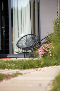 İki dairesel siyah sandalye beyaz perdeli cam bir kapının yanında bir verandada oturuyor. Pembe çiçekler ve bir taş patika yeşillik ile açık hava ayarlarını güçlendirir.