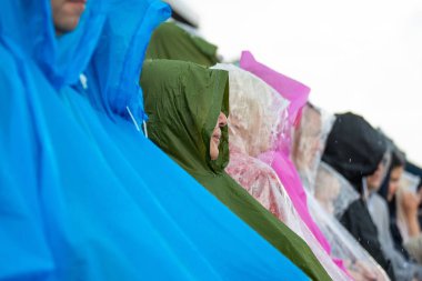 Bir grup birey, mavi, yeşil, pembe ve şeffaf yağmurluk giyip birbirine yakın otururlar. Üzerlerinde görünür yağmur damlaları vardır..