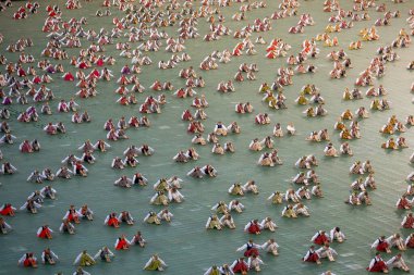 Geleneksel Letonya kostümlü, kırmızı, beyaz ve yeşil desenli bir grup çocuk yeşil bir yüzey üzerinde sıcak ışık altında oturuyorlar..