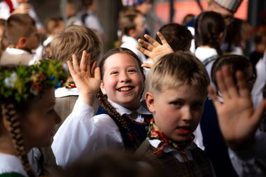 Geleneksel Letonya kostümleri giyen bir grup çocuk. Gülümseyen bir kız merkezde el sallıyor, neşeli yüzler ve canlı detaylarla çevrili..