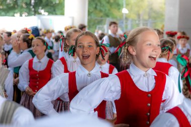 Kırmızı yelekli, beyaz gömlekli ve desenli etekli, renkli kurdelelerle süslenmiş, açık havada ağaçların yanında ve çitlerle çevrili Letonya ulusal kostümleri giyen bir grup çocuk..