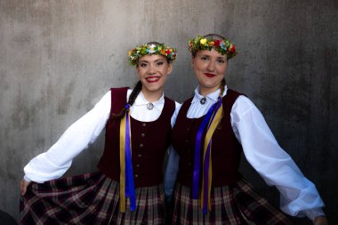 Geleneksel Letonya kostümlü, kestane rengi yelekli, ekose etekli ve çiçekli çelenkli iki genç kız düz beton bir duvara gülümsüyor..