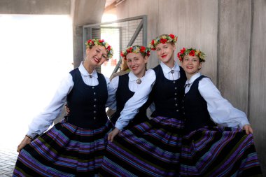 Dört çocuk çizgili etekleri, koyu yelekleri ve çiçekli çelenkleri olan geleneksel Letonya kostümleri giyerler. Beton bir duvar ve metal bir kapının yanında dururlar..