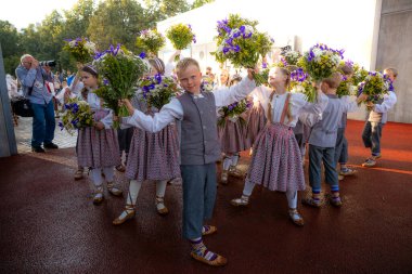 Letonya ulusal kostümlü bir grup çocuk ve kır çiçeği buketleri. Kırmızı asfalt yüzey, modern beyaz yapı ve görünen ağaçlar..