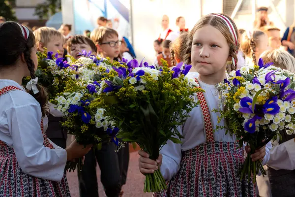 Geleneksel Letonya giysileri içindeki çocuklar kültürel bir toplantı sırasında mavi süsenli, beyaz papatyalı ve sarı aksanlı canlı buketler tutarlar..