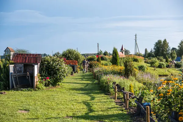 Çiçekler ve ahşap çitlerle çevrili bir bahçe yolu, solunda tavşan kafesleri, kırmızı bir çatı kilisesi, elektrik direkleri ve yürüyen iki kişi..