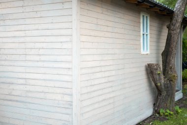 Yatay panelli açık renkli ahşap bir yapı, beyaz çerçeveli bir pencere, koyu kiremitli çatı ve ona yaslanmış bir ağaç gövdesi..