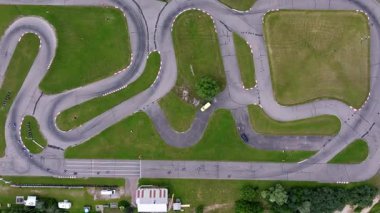 Keskin virajlı bir go kart pisti, çimenli alanlar, küçük bir bina, bir gölet, park edilmiş araçlar ve çerçevede dağınık ağaçlar..