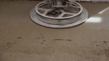 Rotating power trowel blades smooth liquid foam concrete and cement screed floor. Sunlight streams through a window, casting light patterns on the floor.