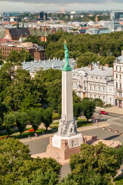 The Monument of Freedom in Riga, Latvia, features a statue holding three stars, surrounded by pathways, trees, historic buildings, and urban activity.