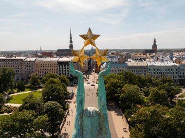 The Monument of Freedom, or Milda, in Riga, Latvia, with three golden stars, urban buildings, St. Peter's Church, trees, pathways, and people visible.