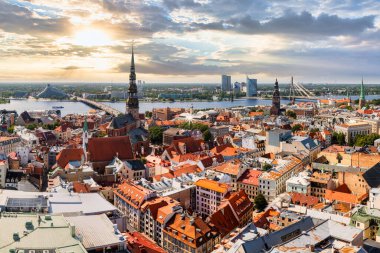 Aerial view of Riga, Latvia, highlighting St. Peter's Church, red roofed buildings, modern bridges, and the Daugava River under warm sunlight.