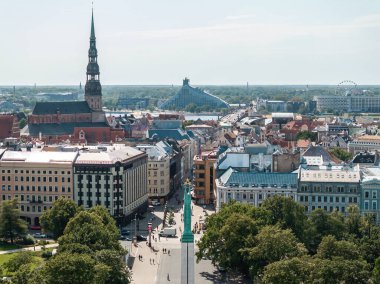 Monument of Freedom in Riga, Latvia, surrounded by trees and pedestrians, with St. Peter's Church, National Library, and Ferris wheel in the background.