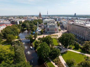 Monument of Freedom in Riga, Latvia, surrounded by a park, pathways, and a canal. Nearby are the Latvian National Opera and Riga TV Tower.