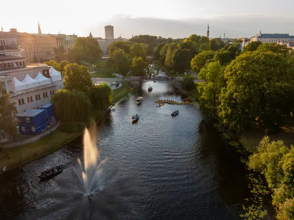 The Monument of Freedom in Riga, Latvia, seen beyond a park with a waterway, boats, a fountain, trees, and urban buildings in warm evening light.