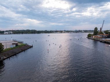 A wide river with paddleboarders scattered across the water, a dock with a crane and industrial structures on the right, and urban elements on the horizon.