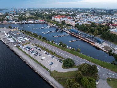 Waterfront area featuring a marina with docks and boats, a road with parking lots, and modern buildings alongside historic red roofed structures.