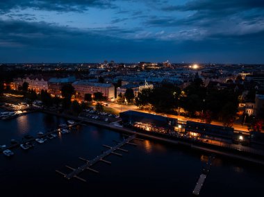 A waterfront marina with docked boats at twilight, illuminated buildings lining the street, and warm lights reflecting on the dark water under an evening sky.