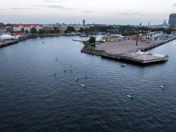A group of paddleboarders on the water near a dock with industrial structures, red roofed buildings, and a cityscape featuring modern and historic architecture.