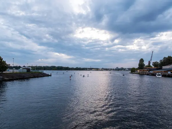 A wide river with paddleboarders, docked boats, and a crane on the right. The left shows a sloped embankment, with buildings and trees on the horizon.
