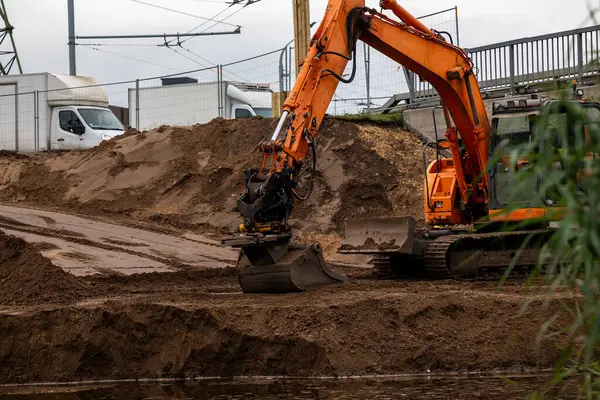 An orange excavator moves soil at a construction site near a body of water, with mounds of dirt, a white van, utility poles, and a bridge visible.