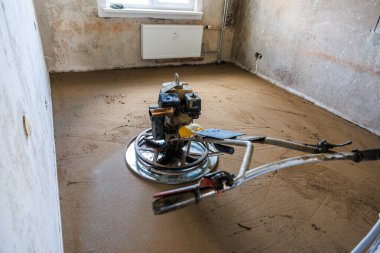 İnşaat alanı tamamlanmamış duvarları olan bir oda, taze beton üzerine döşenmiş bir zemin tarama makinesi, pencerenin altında bir radyatör ve görünür duvar boruları gösteriyor..