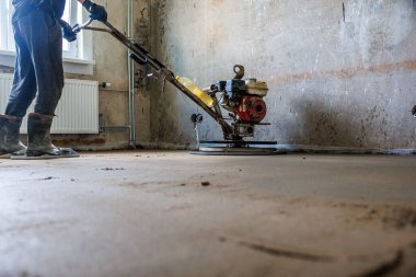 Bir işçi, henüz tamamlanmamış duvarları, radyatörü ve penceresinden gelen doğal ışığı olan bir odada yeni beton bir zeminde güç malası kullanıyor..