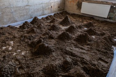 Beton duvarlı bir ev odası, monte edilmiş bir radyatör ve görünürde ayak izleri olan düz olmayan toprak tepecikleriyle kaplı bir zemin..