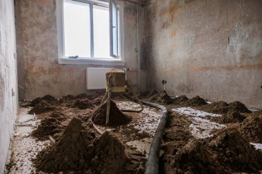 İç mekan, çıplak duvarlar, merkezi bir inşaat aleti, zeminde gevşek toprak ve kısmen açık olan pencerenin altında bir radyatör ile yenileniyor..