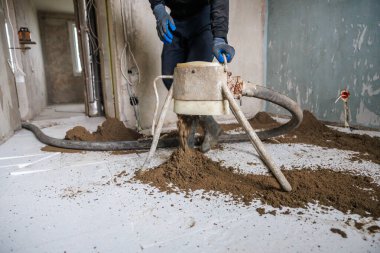 Mavi eldivenli bir işçi, yenilenmiş boruları ve tamamlanmamış duvarları olan bir odanın beyaz fayanslı zemininde bir tarama makinesi kullanıyor..