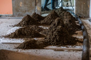 Kapalı bir inşaat alanının beton zemininde birkaç toprak yığını tamamlanmamış duvarlar, açıkta kalmış alçı ve yerde görünür bir boru var..