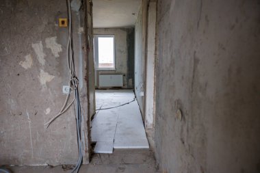 Açık duvarları, görünür kabloları, gevşek döşemeleri ve inşaat malzemeleriyle tamamlanmamış bir iç mekan. Aşağıda radyatör olan bir pencere doğal ışığa izin verir.