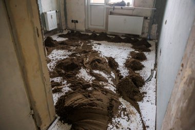 Beyaz fayanslı zeminde gevşek toprak, tamamlanmamış duvarlar, açıkta kalan borular, radyatör ve pencereden içeri giren doğal ışık ile yenilenmiş bir oda..