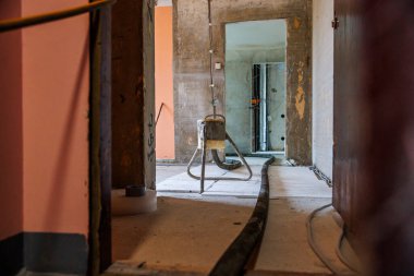 Açık beton duvarları, metal taburesi, yerde siyah kablo, şeftali boyalı bölümler, görünür borular ve kablolar olan kapalı bir inşaat alanı..