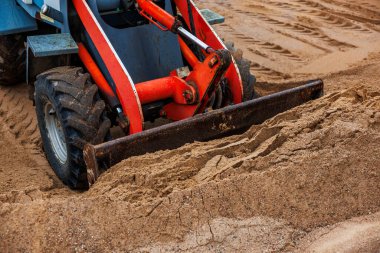 Metal bıçağıyla kum seviyesini yükselten kırmızı ve mavi bir ön yükleyiciye yaklaş. Sert lastikler ve detaylı lastik izleri kumlu şantiyede görülebiliyor..