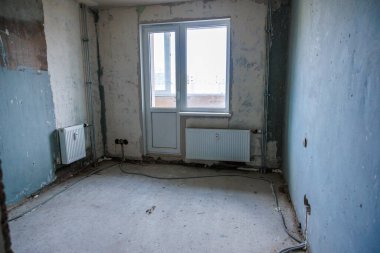 Tamamlanmamış beton döşeme, alçılı duvarlar, açık kablolar, bir pencere, balkona açılan bir kapı ve aşağıya yerleştirilmiş iki radyatörlü bir oda..