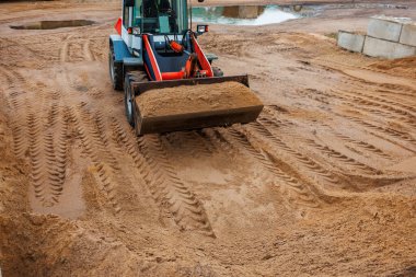 Kırmızı ve siyah aksanlı bir yükleyici inşaat alanında kum taşır. Lastik izleri zemini kaplıyor, beton bloklar ve su dolu bir çukur görünüyor..