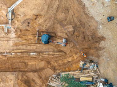 Kazı makinesinin, lastik izlerinin, dağınık malzemelerin, ahşap kalasların, boruların ve köşede küçük bir ağacın olduğu bir inşaat alanının genel görüntüsü..