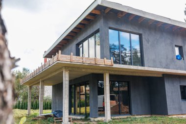 Koyu gri duvarları, büyük camlı pencereleri, düz bir çatısı, açık ahşap kirişleri ve yapım aşamasında olan bir balkonu olan modern iki katlı bir ev..