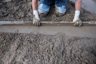 Bir işçi ıslak betonu düzleştirmek için düz bir aletle kum ve çimento yüzeyinde diz çöker. Beyaz eldivenler ve mavi pantolon aktif çalışma belirtileri gösteriyor.