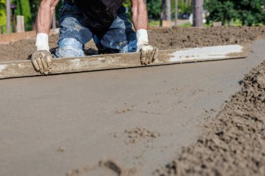 Eldivenli, mavi pantolonlu, boyalı, pürüzsüz, düz bir aletle betonu ıslatan bir işçi. Ağaçlar ve bitkiler uzaktan görülebilir..