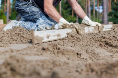 Bir işçi düz bir aletle kumu düzleştirirken, hassasiyete odaklanarak diz çöker. Arka plandaki uzun ağaçlar açık hava ortamını işaret ediyor.