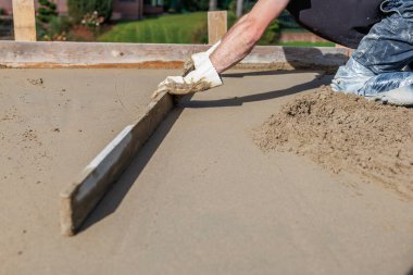 Eldivenli bir işçinin elleri ıslak beton seviyesinde tahta çerçeveli bir yüzey üzerinde düz bir alet kullanarak bulanık konut yapıları ve bitki örtüsüyle görülebilir..