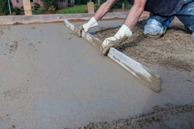 Beyaz eldivenli ve mavi kotlu bir işçi düz bir aletle ıslak betonu düzleştirirken diz çöker. Arka planda bir tuğla bina ve bitki örtüsü görünür..